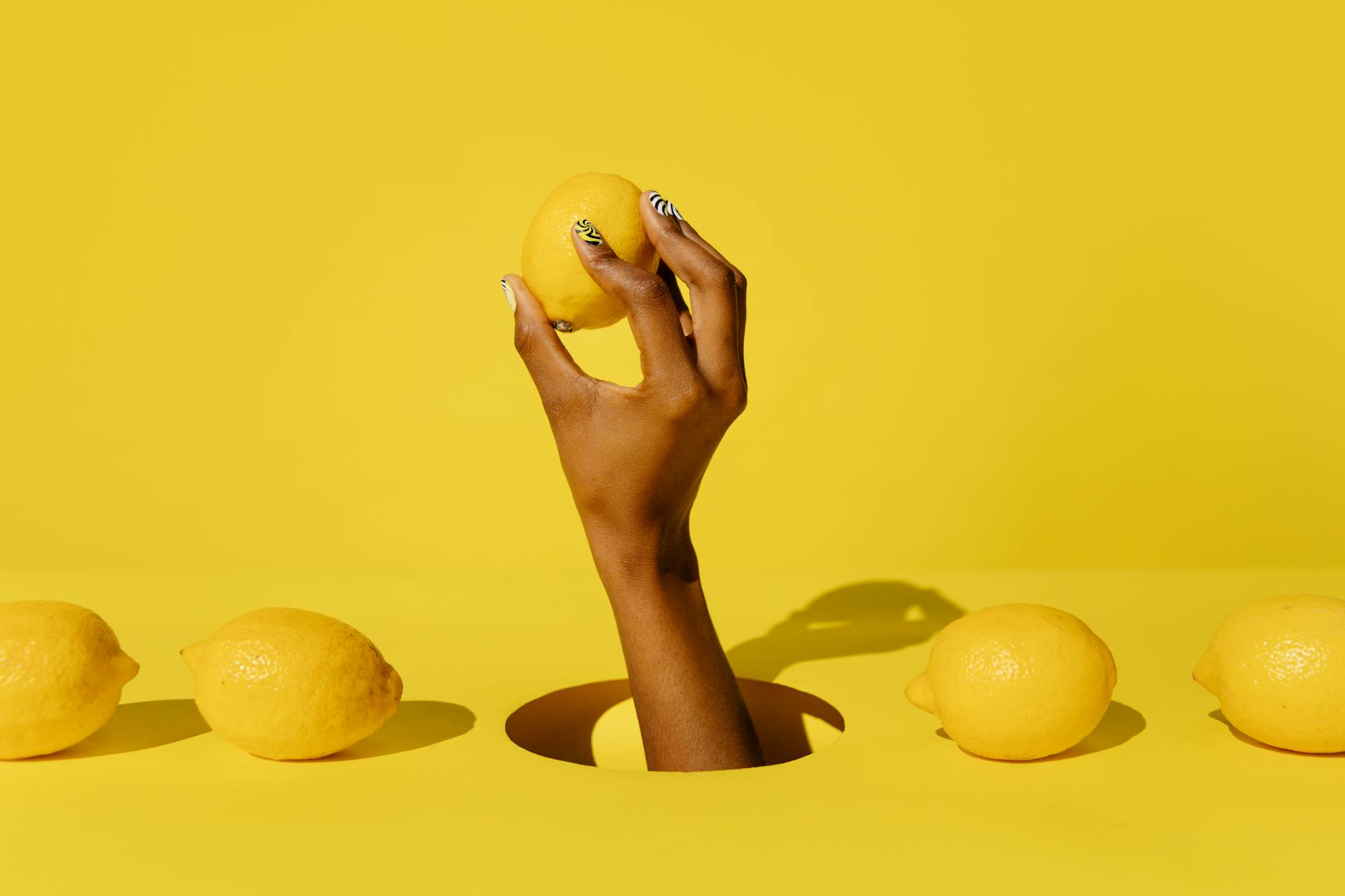 A hand holding a bright yellow lemon against a vivid yellow background, representing the fresh citrus design of the Lem vibrator